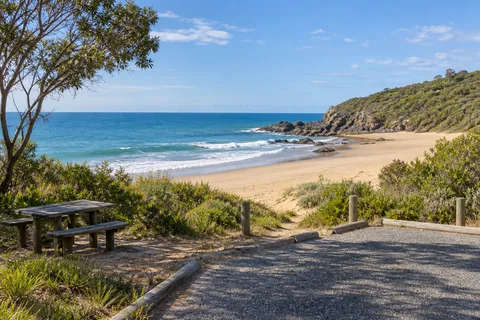 AI-generated editorial illustration of a vegetated coastal access area with a picnic table, gravel parking and a sandy beach near a rocky headland.